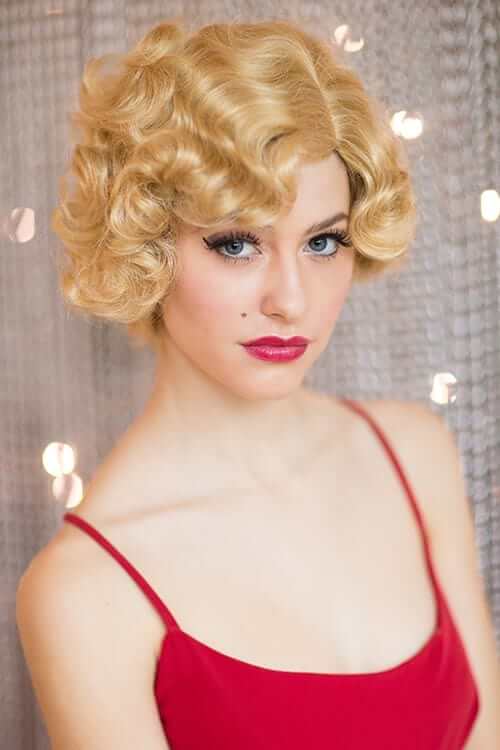 A woman wearing a blonde wig with short, finger wave curls and an off-center parting. Blonde 1920s 1930s style wig, short with finger waves: Diva warm blonde 24B