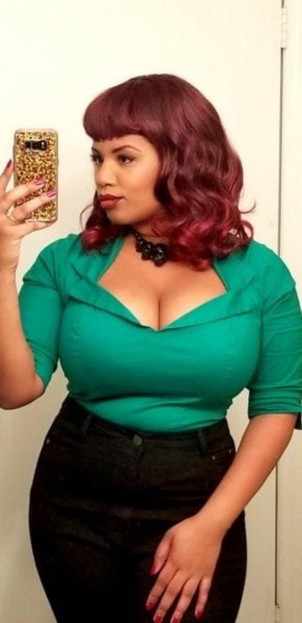 Woman in a green top and black pants holding a phone with a leopard print case. Black and red pinup style wig, gently wavy with short fringe, 1950s style: Lillian