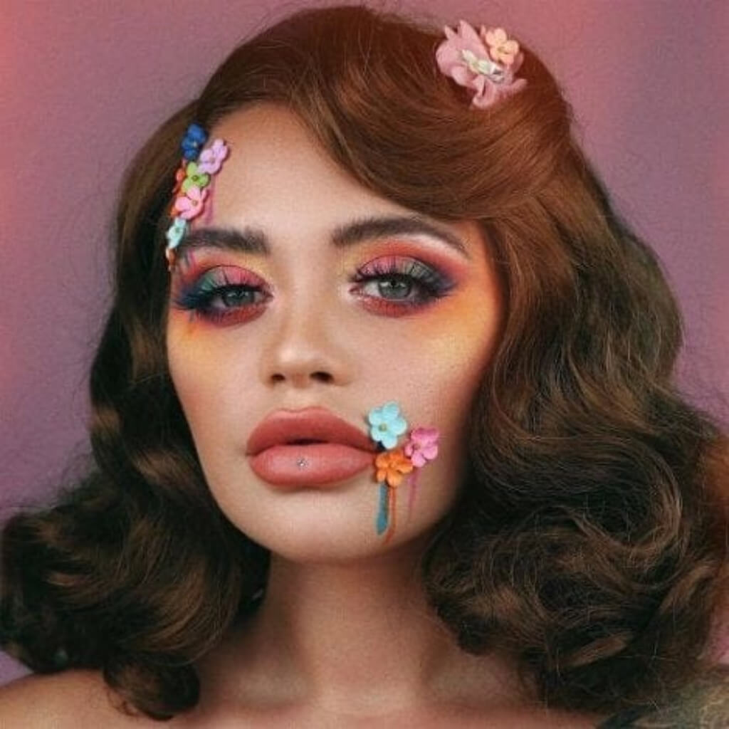 Woman with colourful makeup and floral hair accessories against a pink background. Brown 1940s pinup style wig, with finger waves: Honey light brown 12