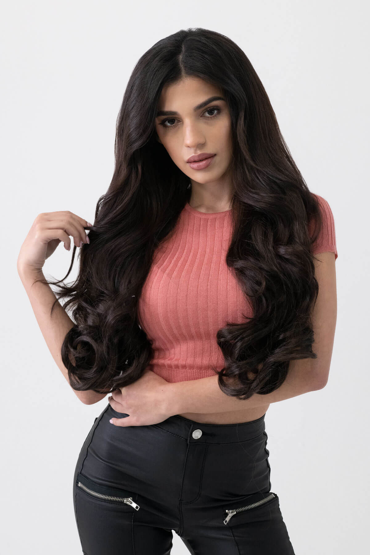 Woman with long, wavy hair wearing a pink top and black pants on a white background