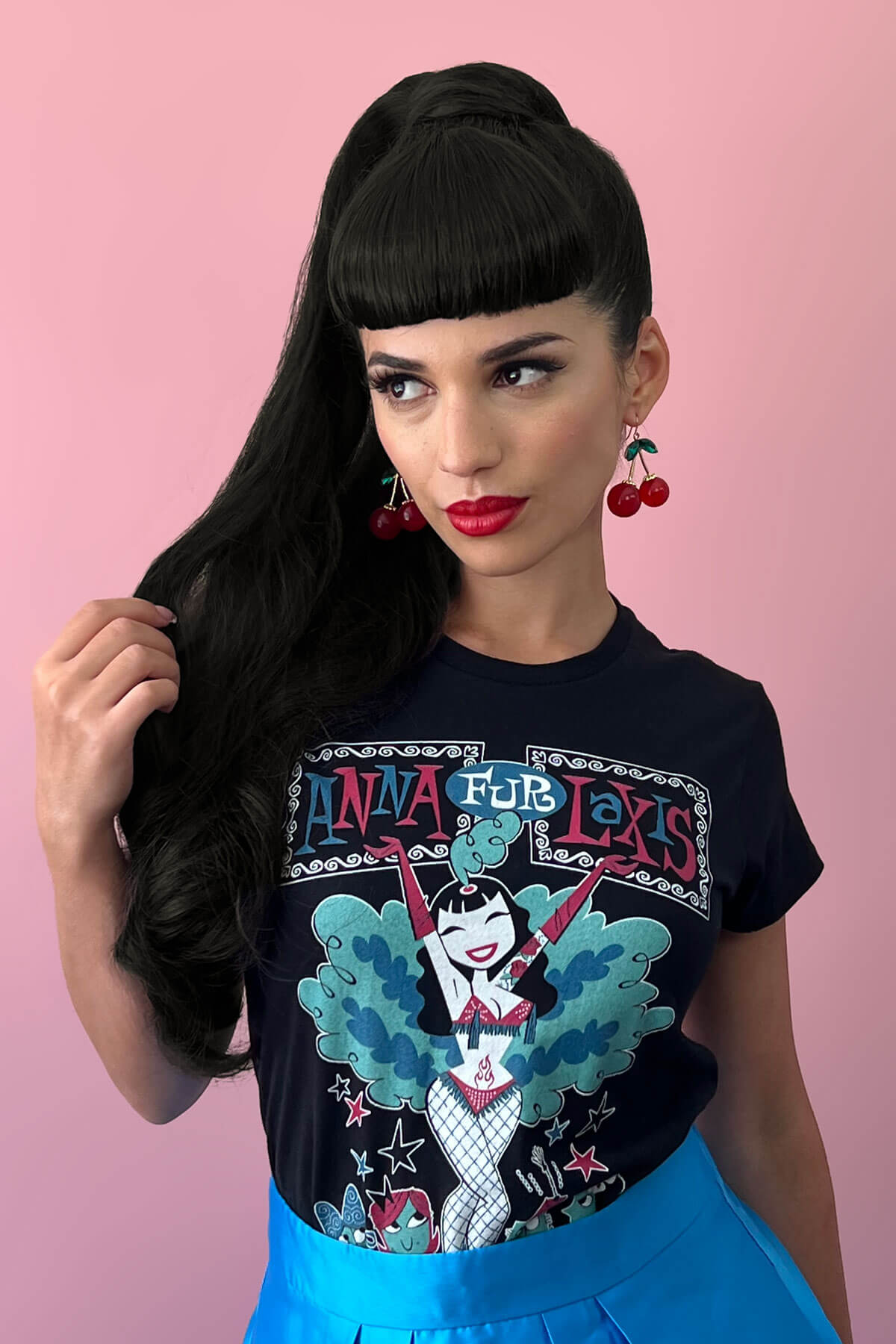 Woman wearing a black t-shirt with colorful graphic design against a pink background. Long ponytail extension with soft waves: Sophie black 1b