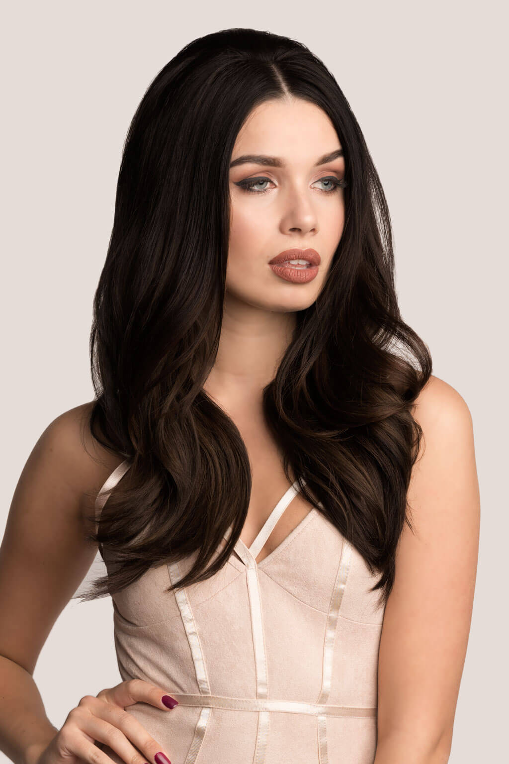 Woman with long, dark brown hair wearing a beige dress against a light gray background. Volume and Height Booster Hairpiece: Blaise - champagne blonde 613