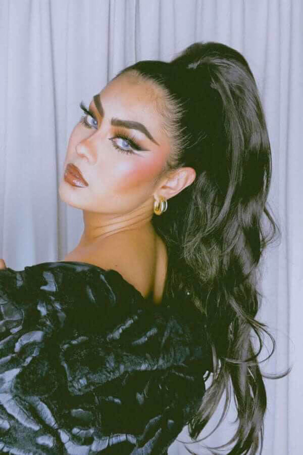 Woman with styled hair and makeup wearing a black textured garment against a light background