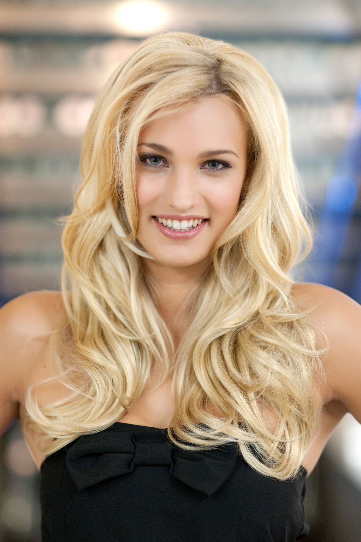 Woman with long blonde hair wearing a black top against a blurred indoor background. Heat styleable half wig hairpiece (3/4 wig), long, wavy, Flexihair: Michela blonde 24BH613