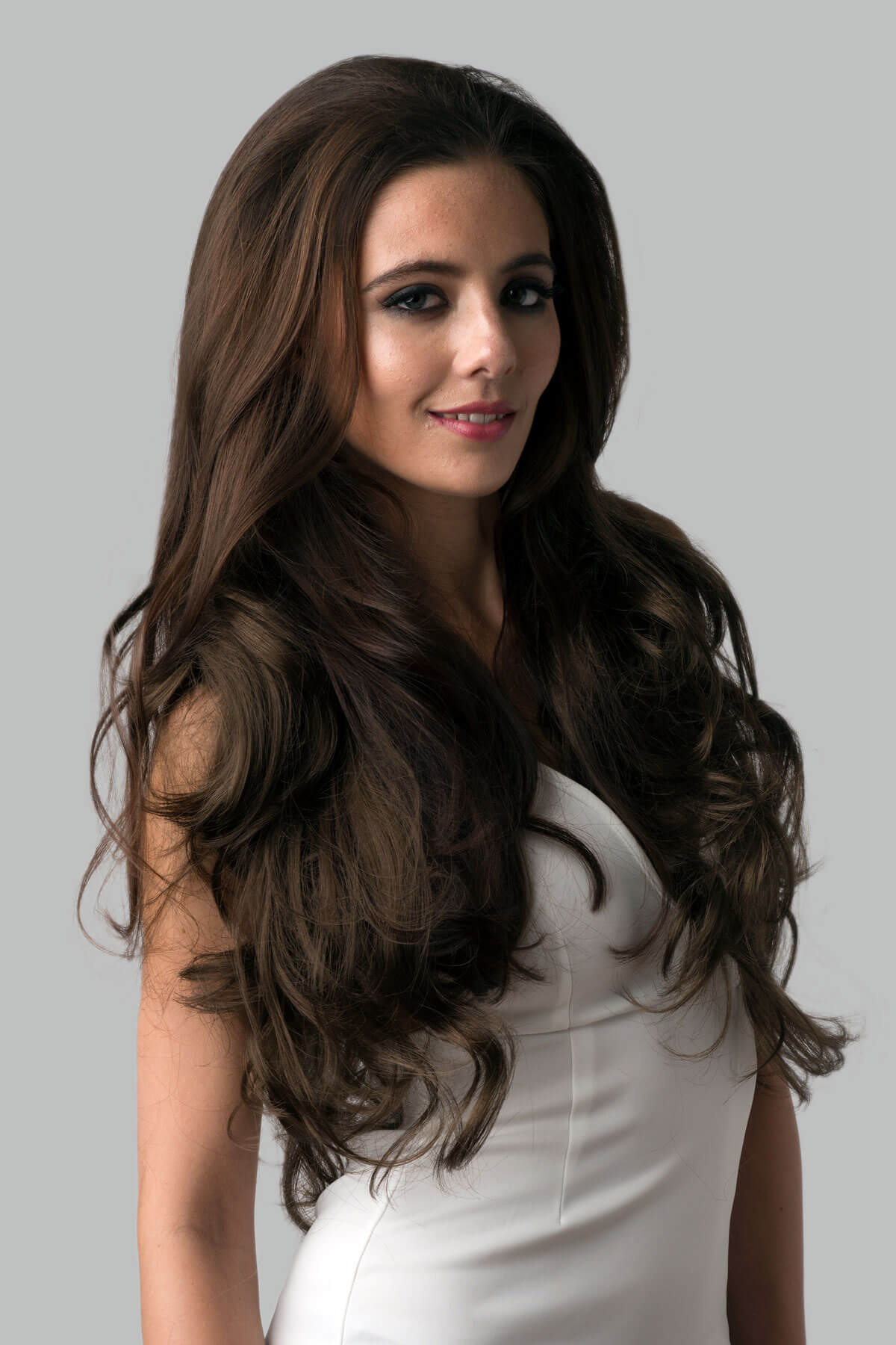 Woman with long, wavy brown hair wearing a white dress against a gray background.  Brown half wig hairpiece, full, long with gentle waves: Taya mocha brown 10