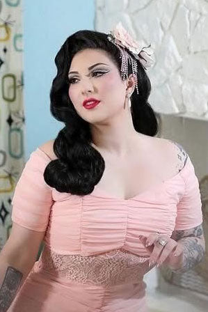 Woman in a pink dress with a decorative headpiece against a light blue wall. 1950s style half wig hairpiece with beautiful waves: Morgana black 1B