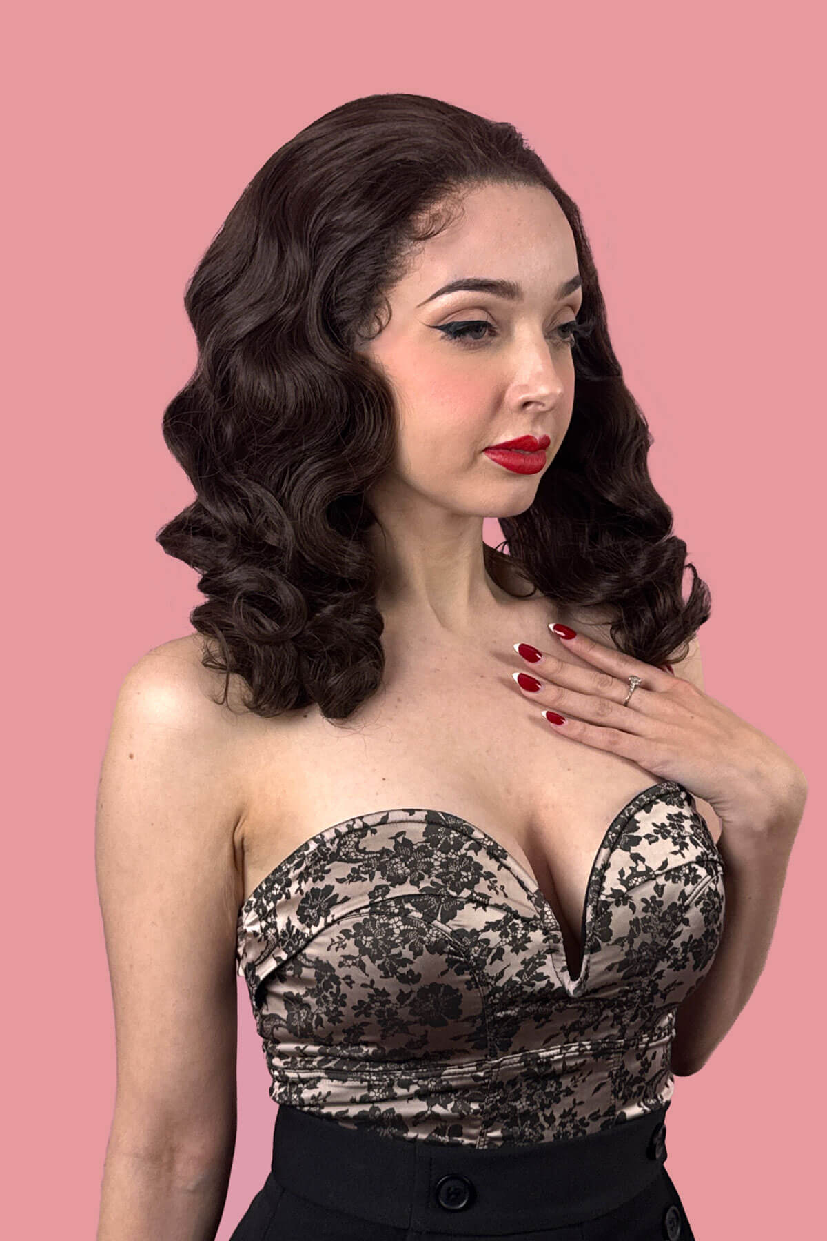 Woman wearing a black lace strapless top against a pink background. Brown pinup wig, lace front with finger waves: Nikita