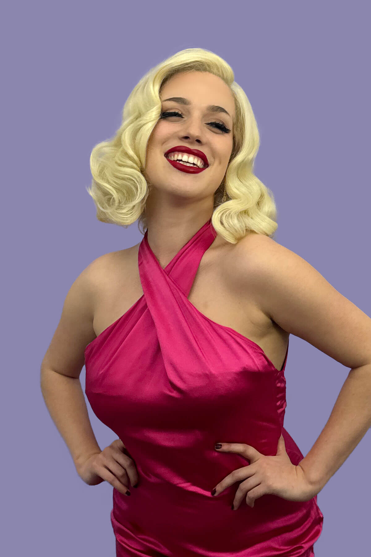 Woman in a pink dress with a purple background. Blonde lacefront wig, pinup/vintage style, mid length with finger waves: Della creamy blonde 613