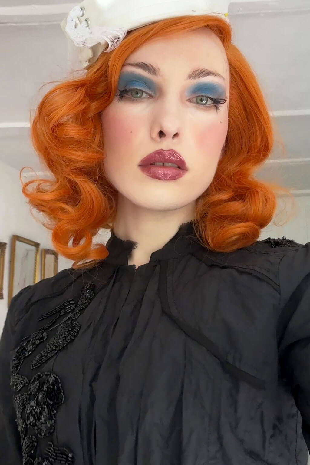 Person with red hair and vintage makeup wearing a black outfit with floral details. Ginger lacefront wig, pinup/vintage style, mid length with finger waves: Flora