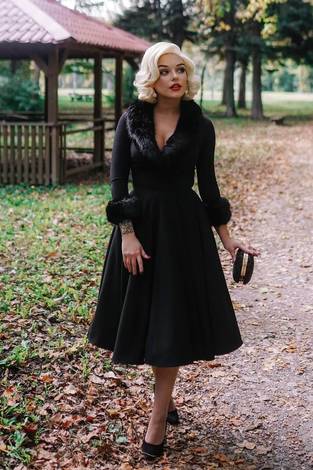 Woman in a black dress with fur trim standing on a path in a park. Blonde pinup wig, lace front, vintage style: Marilyn white blonde 1001