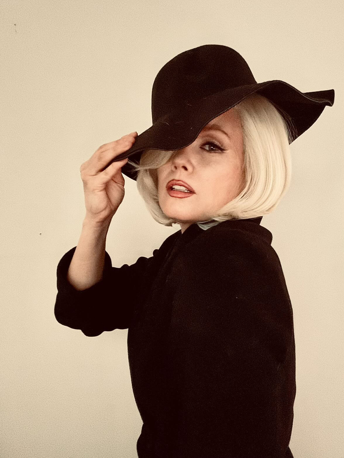 A woman with a short platinum blonde bob wears a black wide-brim hat and a black top against a plain beige studio background. Blonde lacefront wig, pinup/vintage style, mid length, hollywood elegance: Norma Jeane