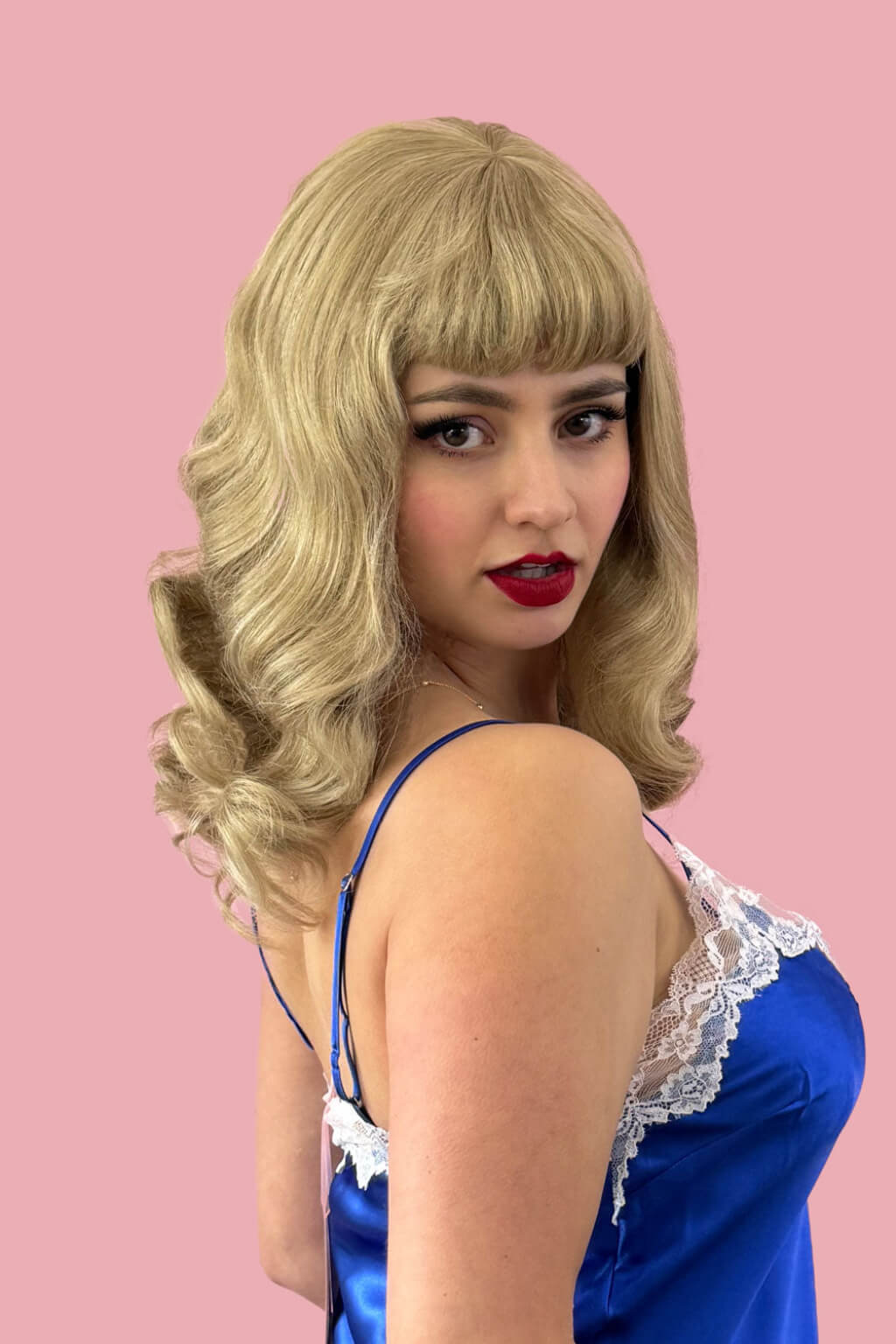 Woman wearing a blonde wig and blue satin top with lace trim against a pink background. Blonde pinup wig, curled with short fringe, 1950s style: Cora blonde 24H613