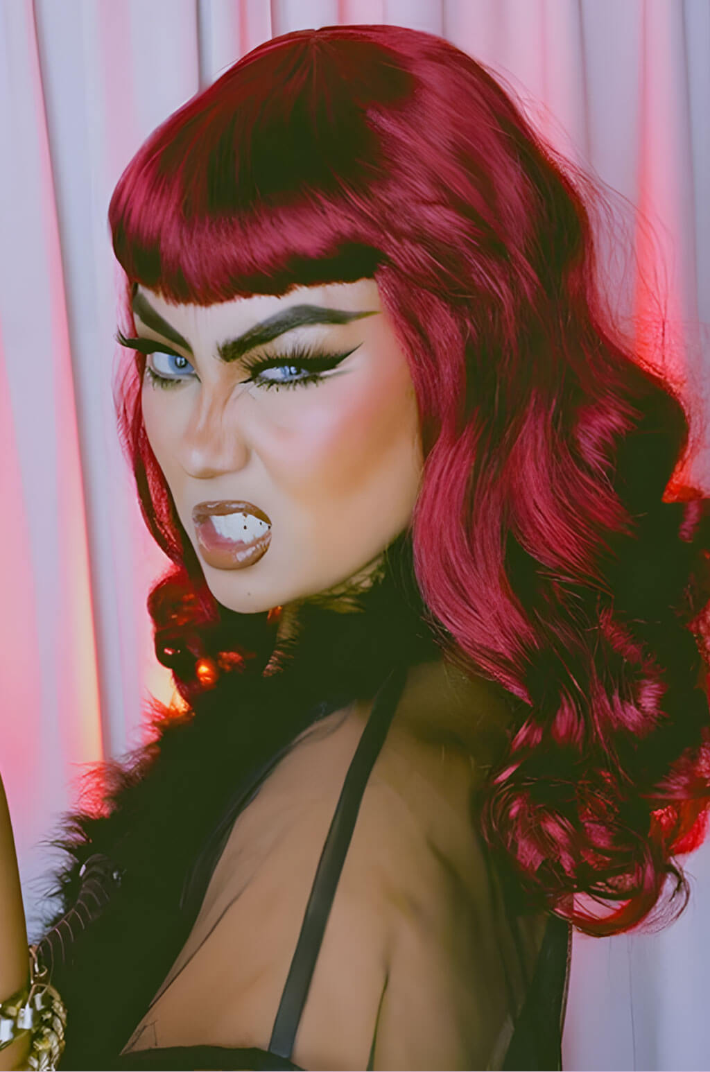 Person with vibrant red hair and dramatic makeup against a light curtain background. Cherry red 1950s pinup style wig: Sandra