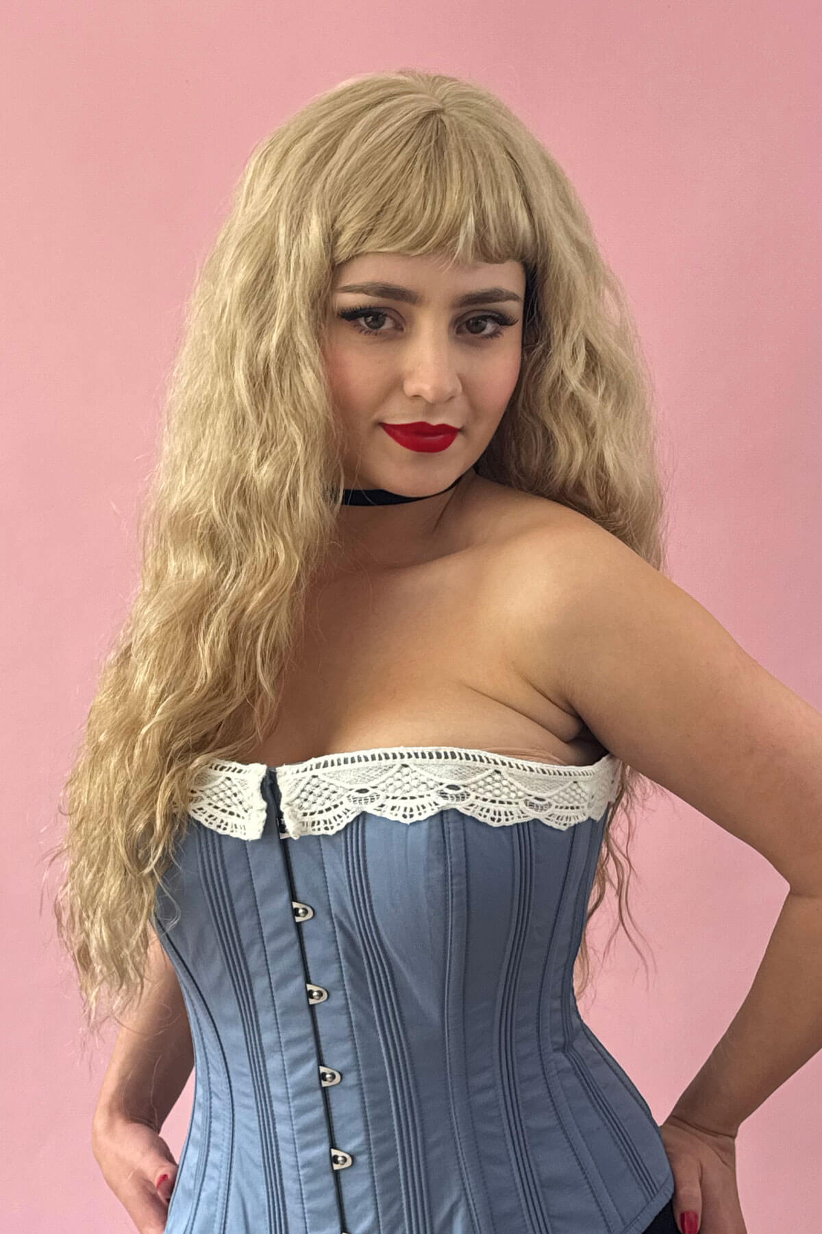 Woman wearing a blue corset with white lace trim against a pink background. Long wig with crimped mermaid waves: Astrid