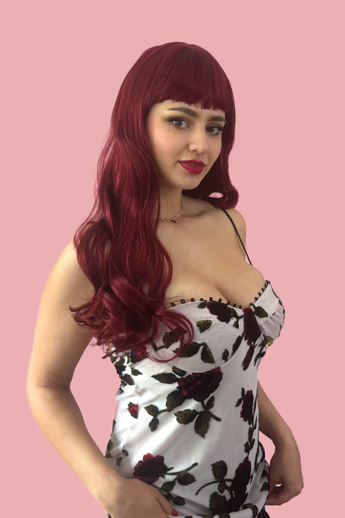 Woman with long red wig wearing a strapless floral dress against a pink background. Long red wig with straight retro-style fringe: Esther