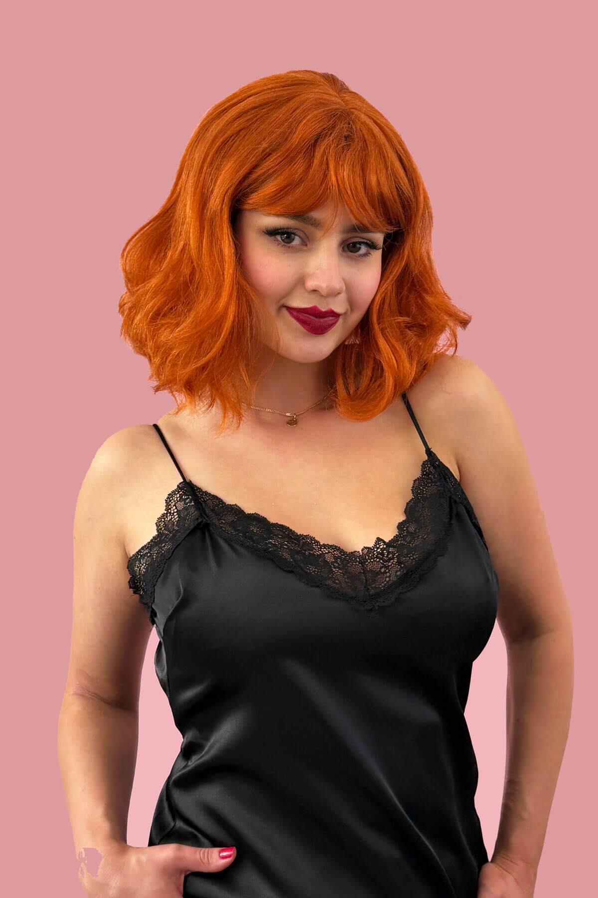Woman with bright orange hair wearing a black satin slip against a pink background. Ginger long wavy bob (lob) wig with swept fringe: Rusty
