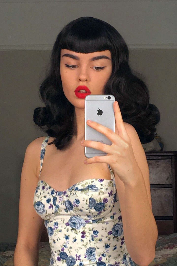 Woman with black hair and red lipstick taking a mirror selfie, wearing a floral dress. Black pinup style wig, curled with short fringe, 1950s style: Bettie 1B