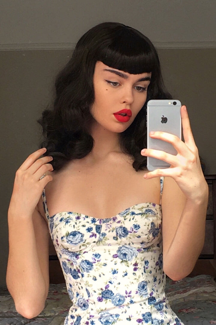 Woman in a floral dress taking a mirror selfie with a smartphone. Black pinup style wig, curled with short fringe, 1950s style: Bettie 1B
