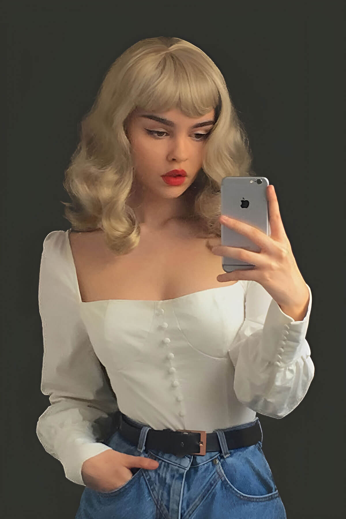 Woman taking a selfie with a smartphone against a dark background. Blonde pinup wig, curled with short fringe, 1950s style: Cora blonde 24H613