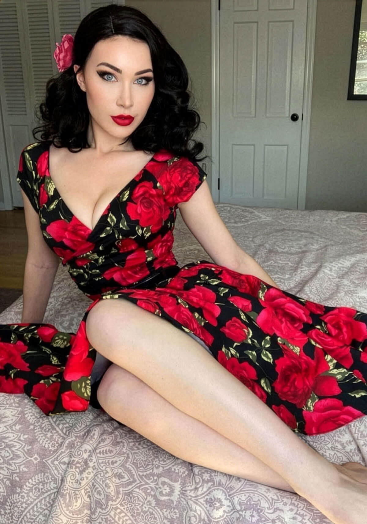 Woman wearing a red and black floral dress sitting on a bed. Black 40s style wig with beautiful marcel waves: Dita