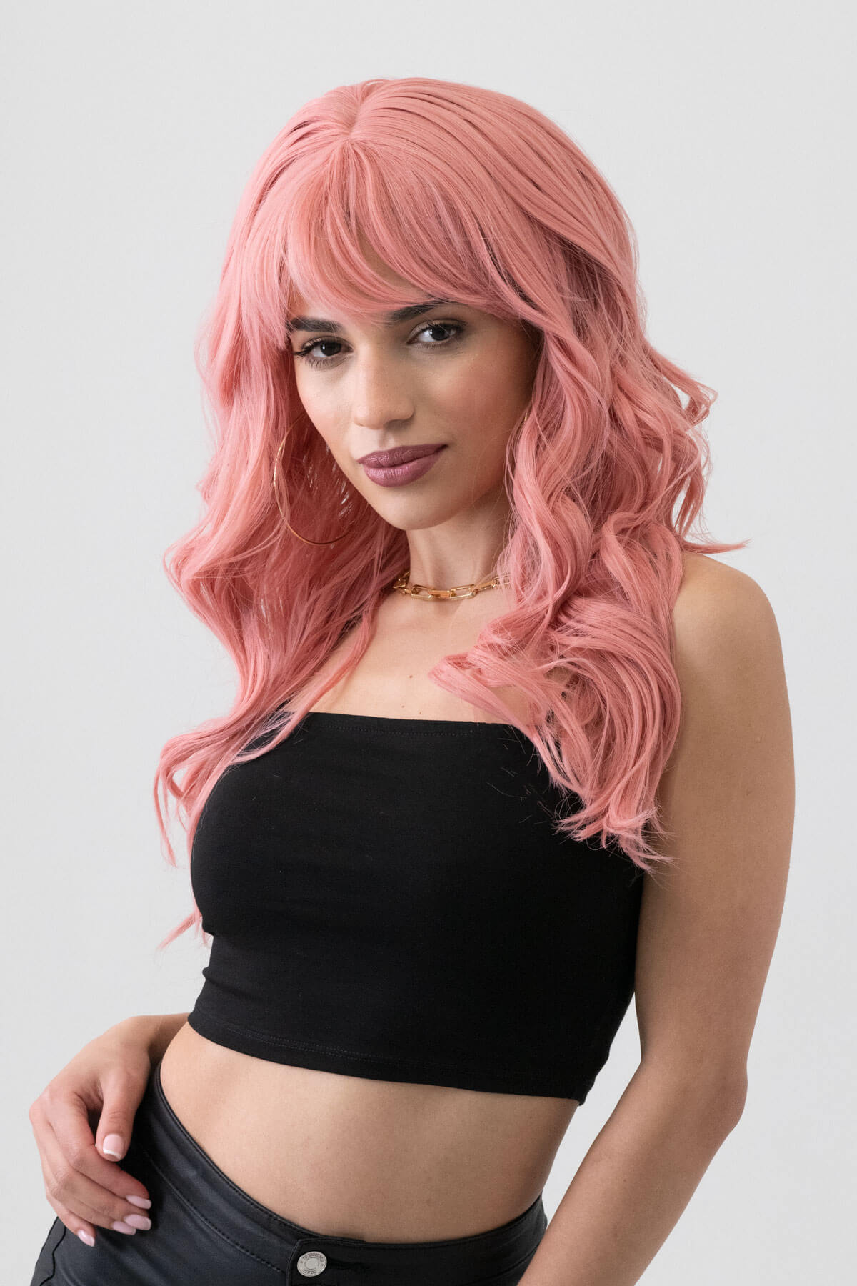 Woman with pink hair wearing a black top against a white background. Dusty pastel pink wig with long cascading waves: Rosalie