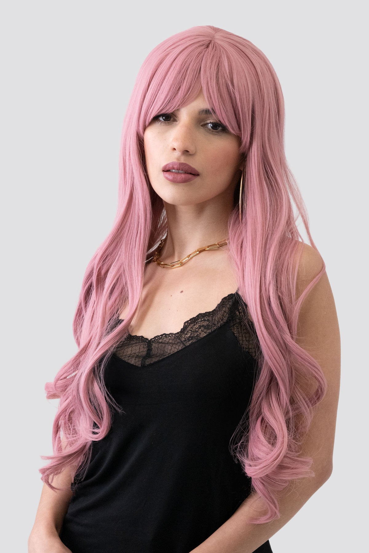 Woman with long pink wig wearing a black top on a light gray background. Long wavy pink wig with sweeping fringe: Millicent