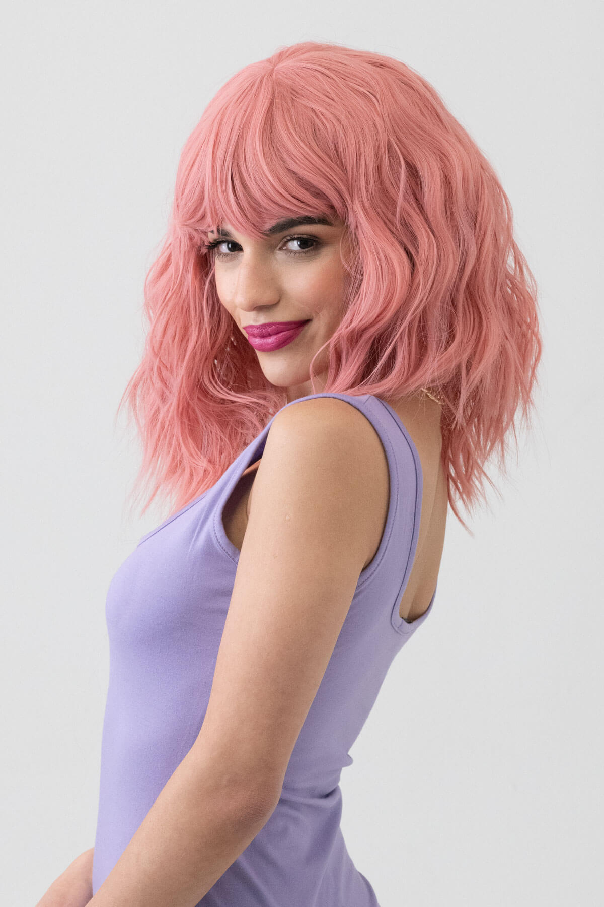 Woman with pink wig wearing a purple dress against a light gray background. Dusty pink long wavy bob wig (LOB) with textured waves: Fearne