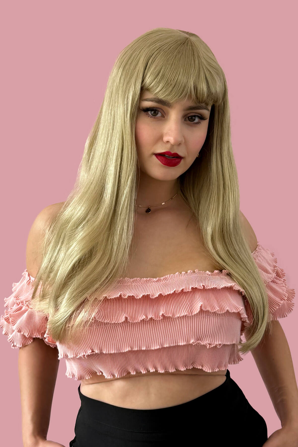 Woman wearing a pink off-shoulder top with ruffles against a pink background. Long blonde wig with straight hair and a short, straight fringe: Nicole ash blonde 24