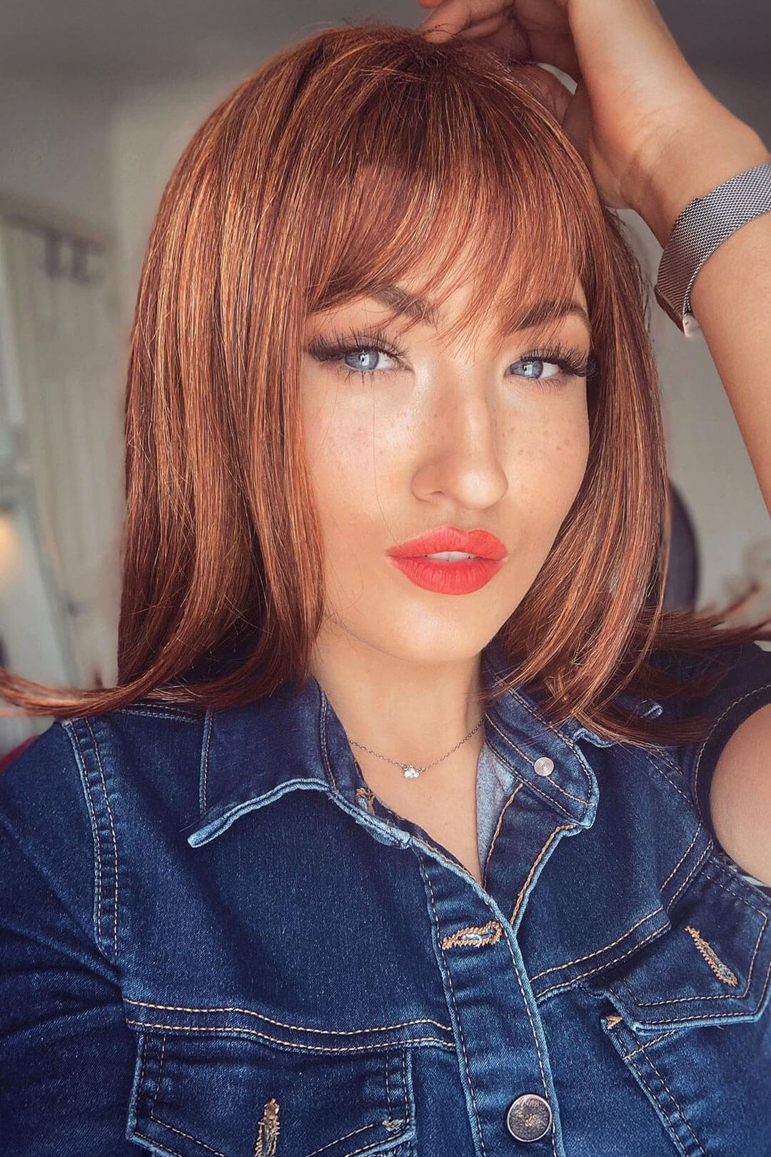 Woman with styled auburn  hair and denim jacket in a blurred indoor setting. Gingery auburn wig with blonde highlights face frame style: Geri