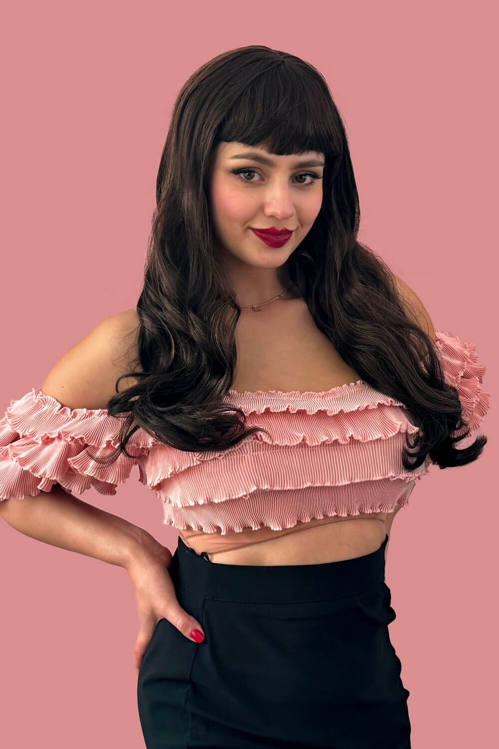 Woman wearing a pink ruffled top against a pink background. Long chestnut brown wig with blonde highlights and pinup-style fringe: Zola
