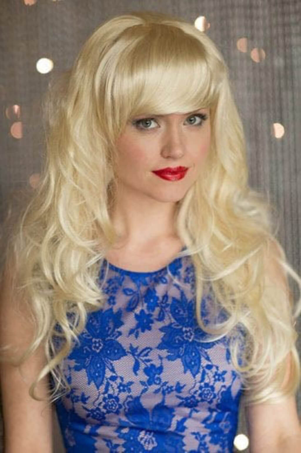 Woman with blonde wig and red lipstick wearing a blue lace dress against a neutral background.  Clip-in fringe hairpiece, curved, side-sweeping/clip-in bangs: Eliana blonde 24BH613 