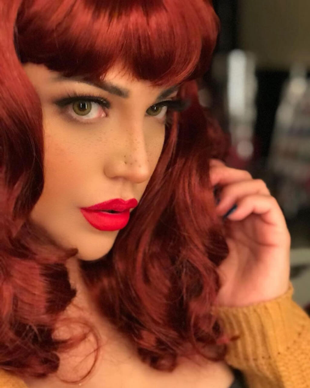 Woman with vibrant red hair and red lipstick posing indoors. Copper red 1950s pinup wig, curled with short fringe: Kitty